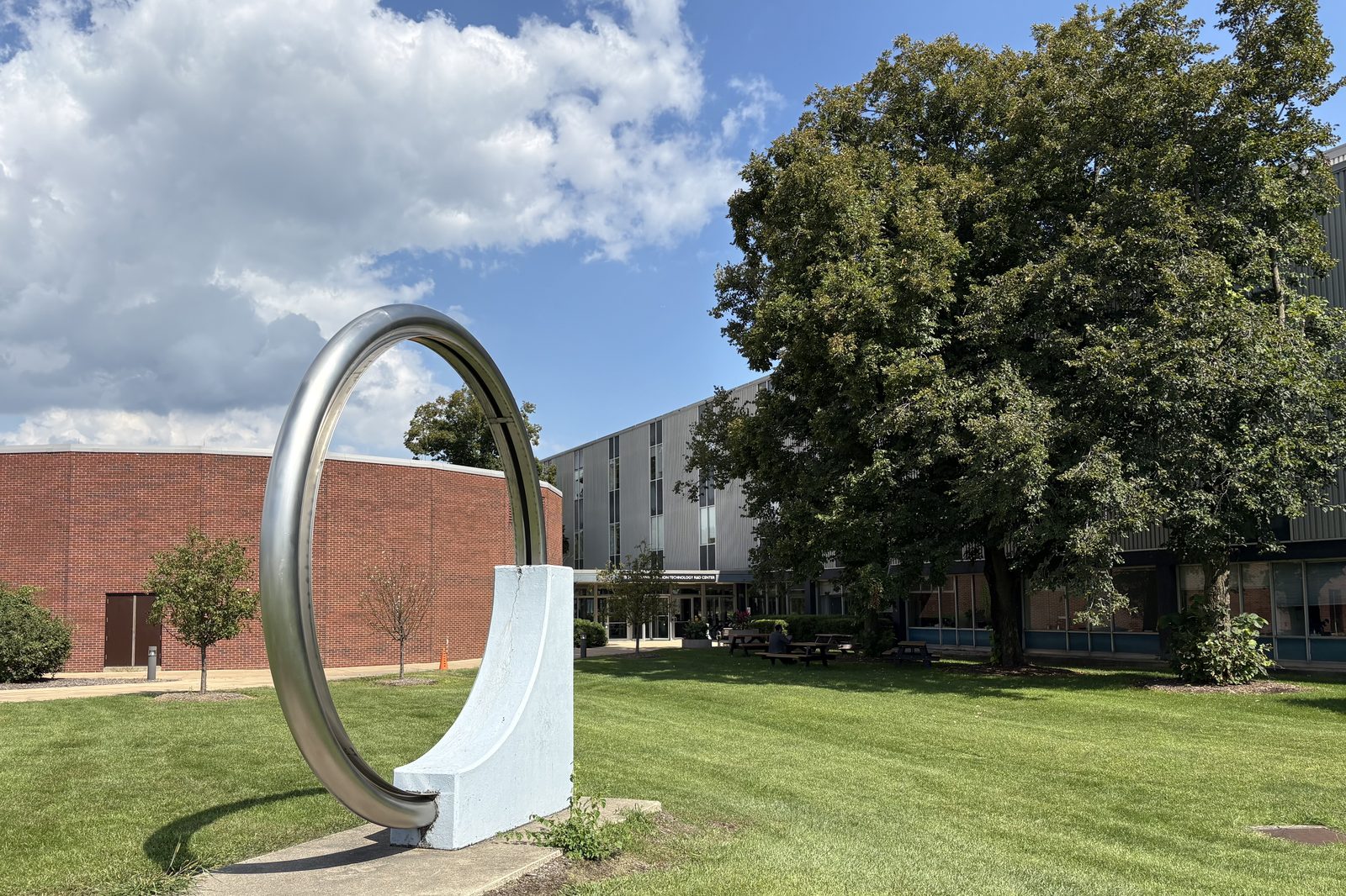 A large steel ring sculpture on the lawn outside Building 362 at Argonne National Laboratory, with the building and a mature oak tree behind it.